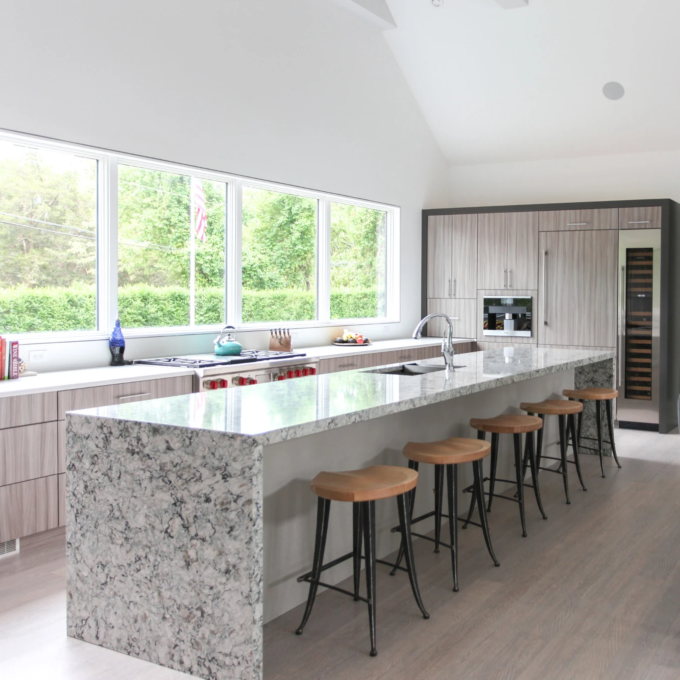 an open and airy kitchen with stunning waterfall-edge island topped with Cambria Praa Sands, a modern glass dining table with plenty of seating, and large windows overlooking a lush garden.