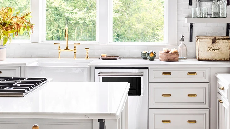 a white kitchen with a porcelain farmhouse sink, brass fixtures, and Ella quartz countertops
