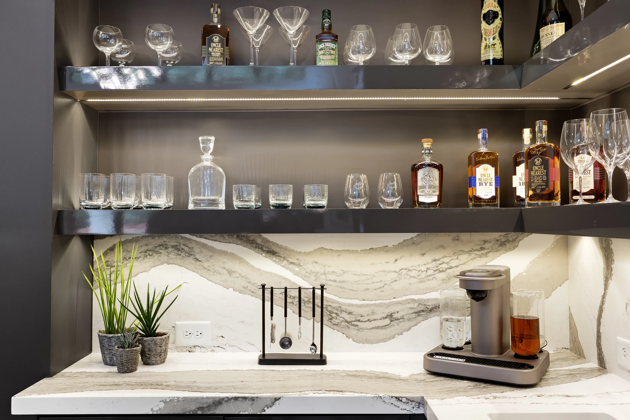 a wet bar with quartz countertops and backsplash, black cabinets, black shelving with LED under-lighting, a coffee machine, and various alcoholic beverages on the shelves.
