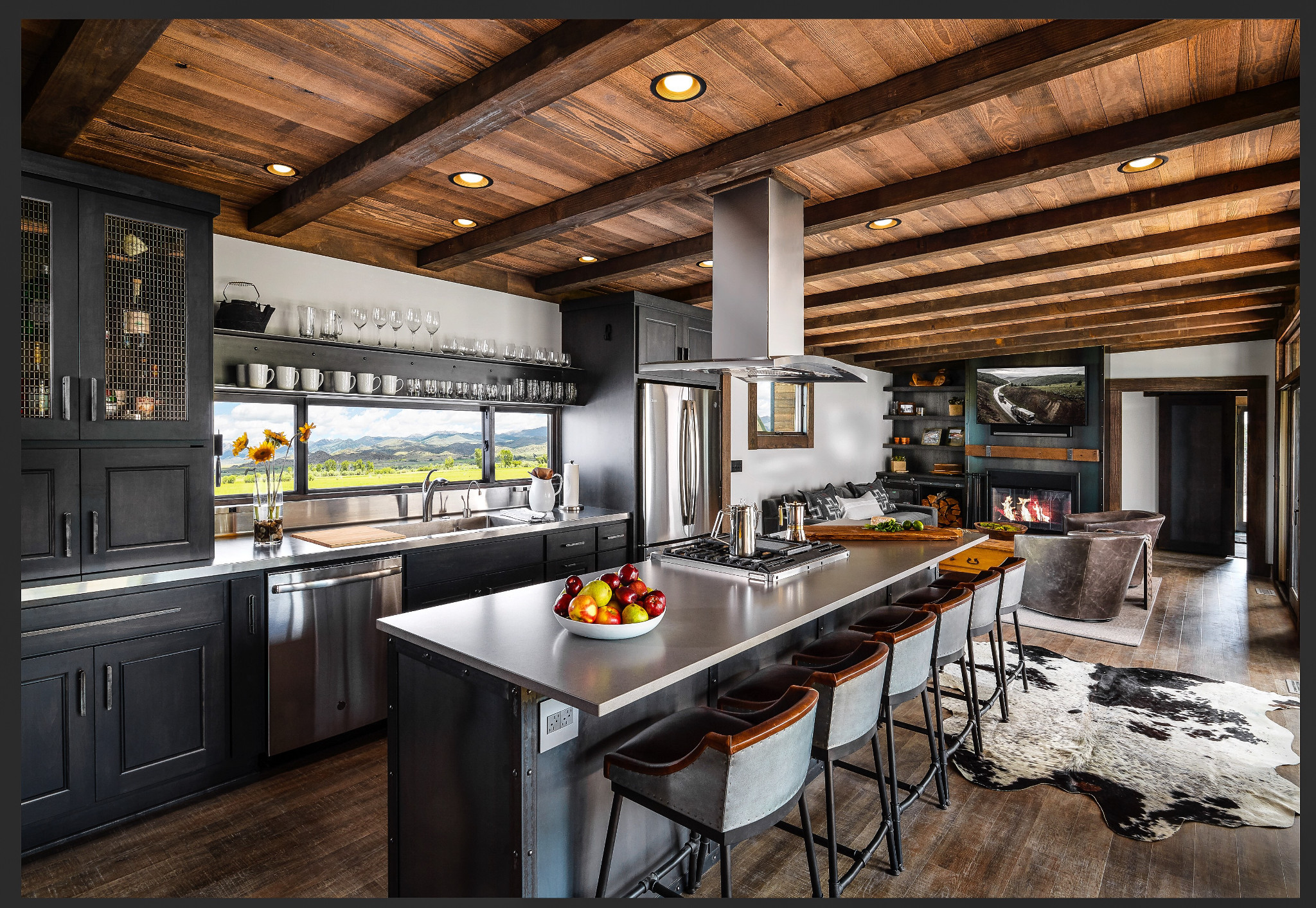 a rustic industrial kitchen next to a cozy living space, with black cabinets, a window above the sink that frames a scenic view, and a stove and range built into the island that is topped with Cambria Devon Matte quartz countertops.