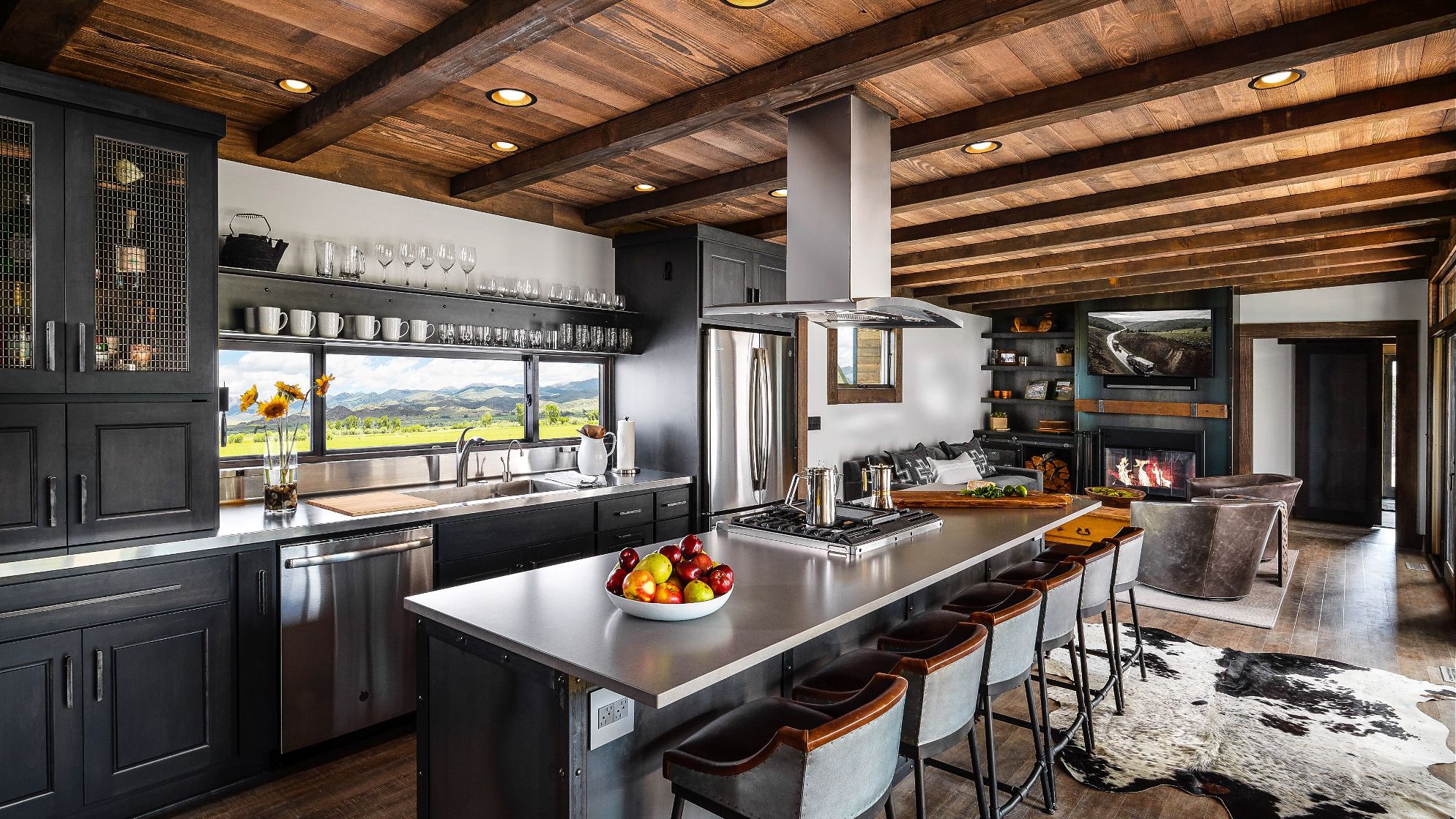 a rustic industrial kitchen next to a cozy living space, with black cabinets, a window above the sink that frames a scenic view, and a stove and range built into the island that is topped with Cambria Devon Matte quartz countertops.