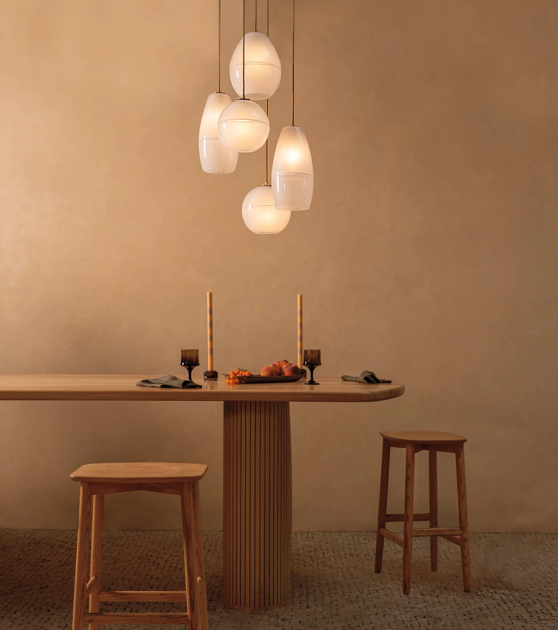 a modern wooden dining table with two wooden stools placed at the front and side, with brown flooring and walls, and 5 blown glass bulbs hanging above, all creating a warm and inviting space. 