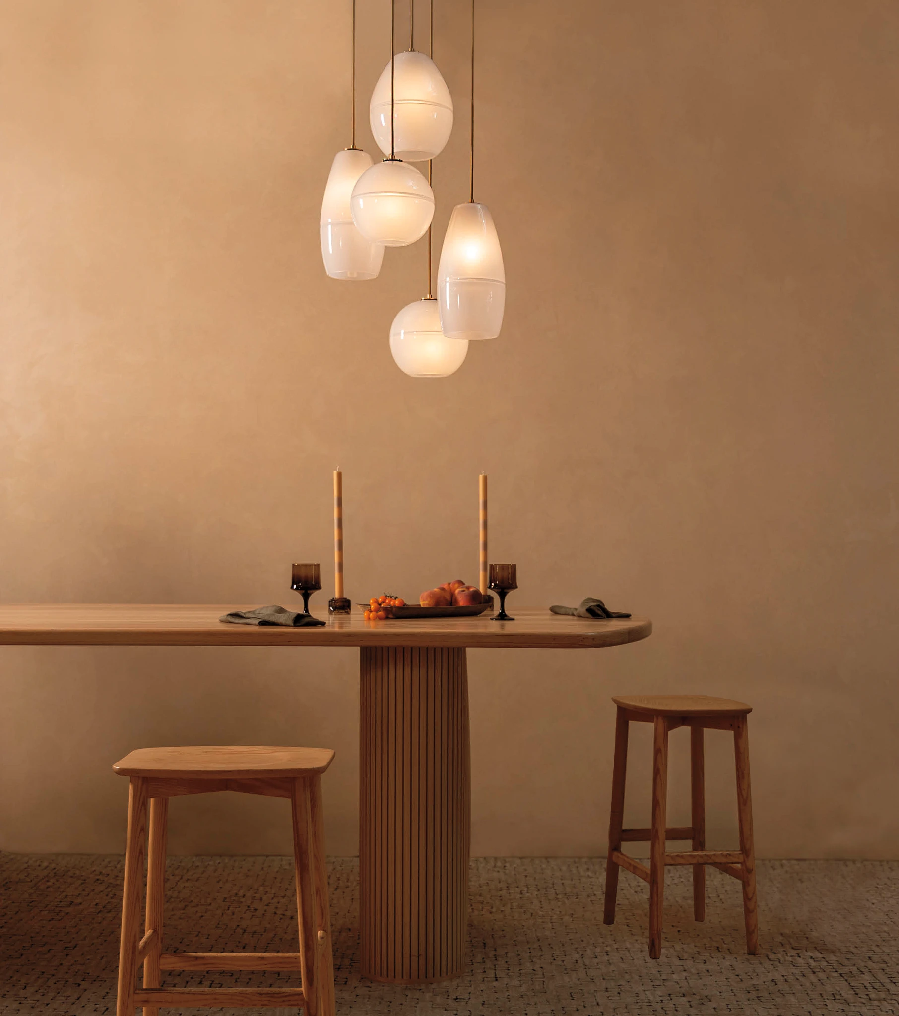 a modern wooden dining table with two wooden stools placed at the front and side, with brown flooring and walls, and 5 blown glass bulbs hanging above, all creating a warm and inviting space.