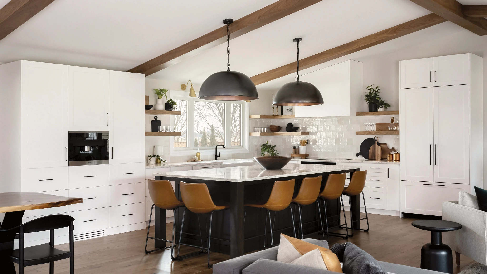 a warm and white kitchen with white cabinets, open wooden shelving, vaulted ceilings with wooden beams, and a black center island topped with white quartz countertops and 6 wooden and black bar stools surrounding it.